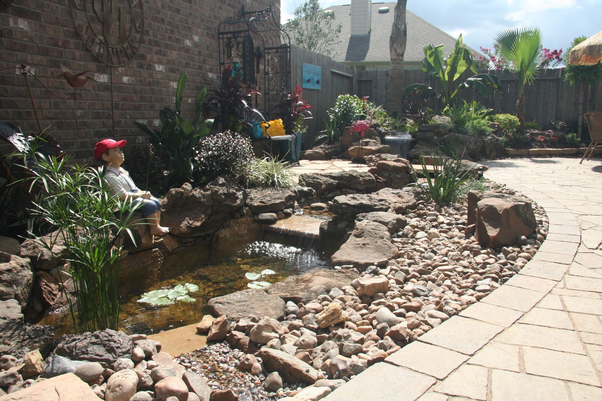 Stone garden path beside a small pond with rocks, flowers, and potted plants in bright sunlight