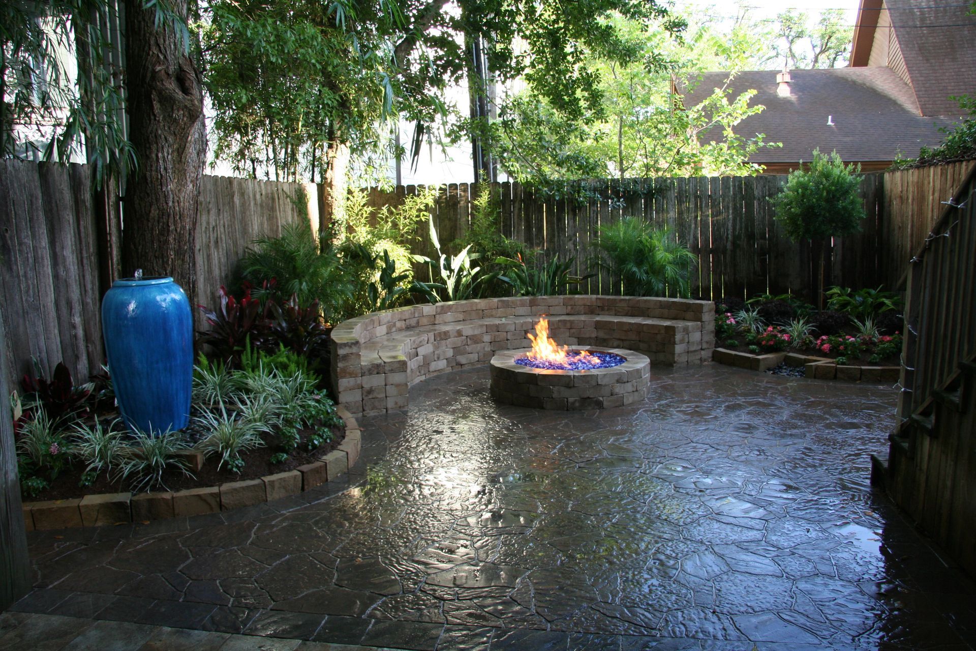 Rainy backyard patio with a lit fire pit, stone wall, trees, and a blue rain barrel