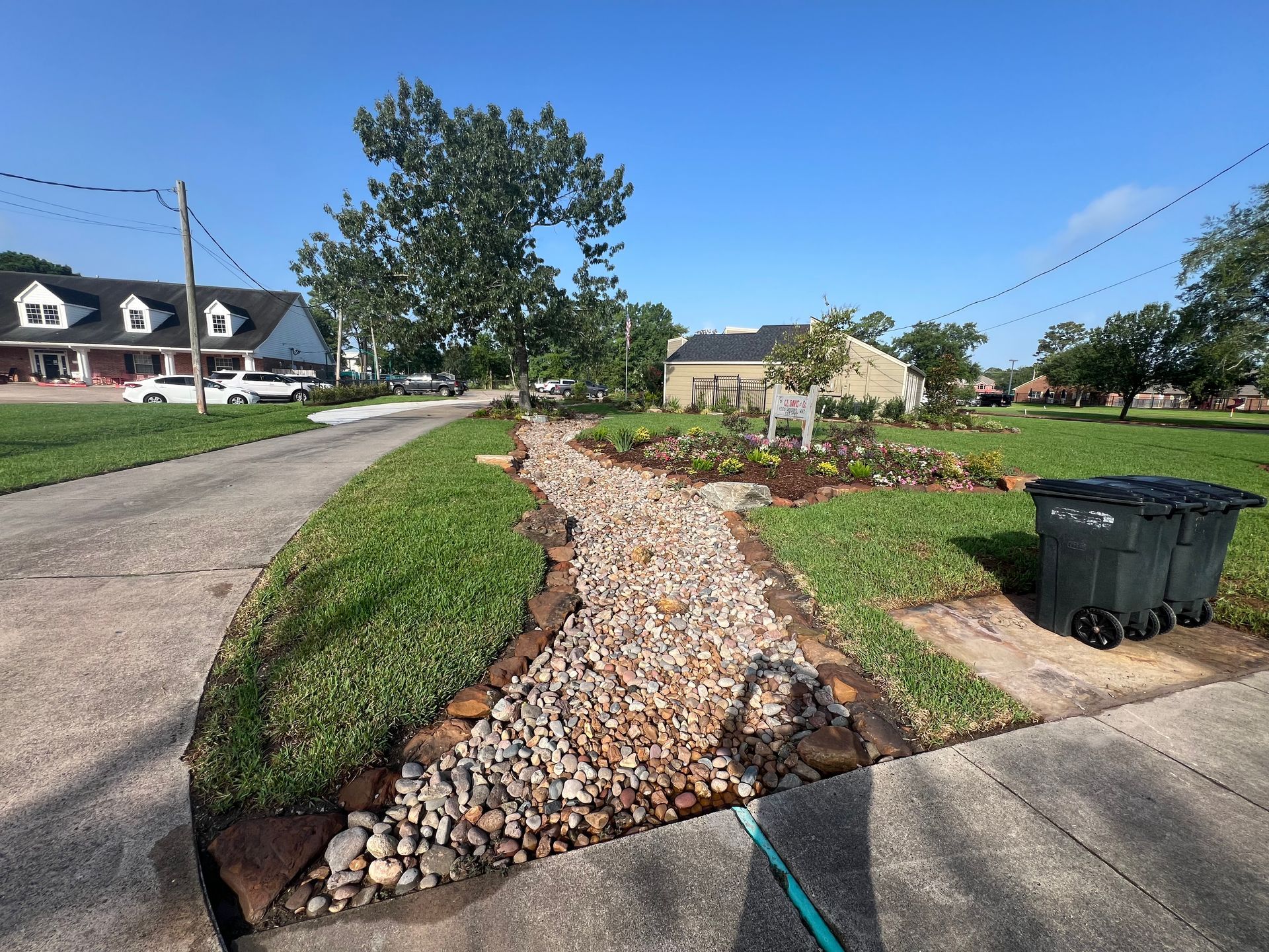Rock-lined drainage swale beside a residential street, with green grass, houses, and trash bins under a blue sky