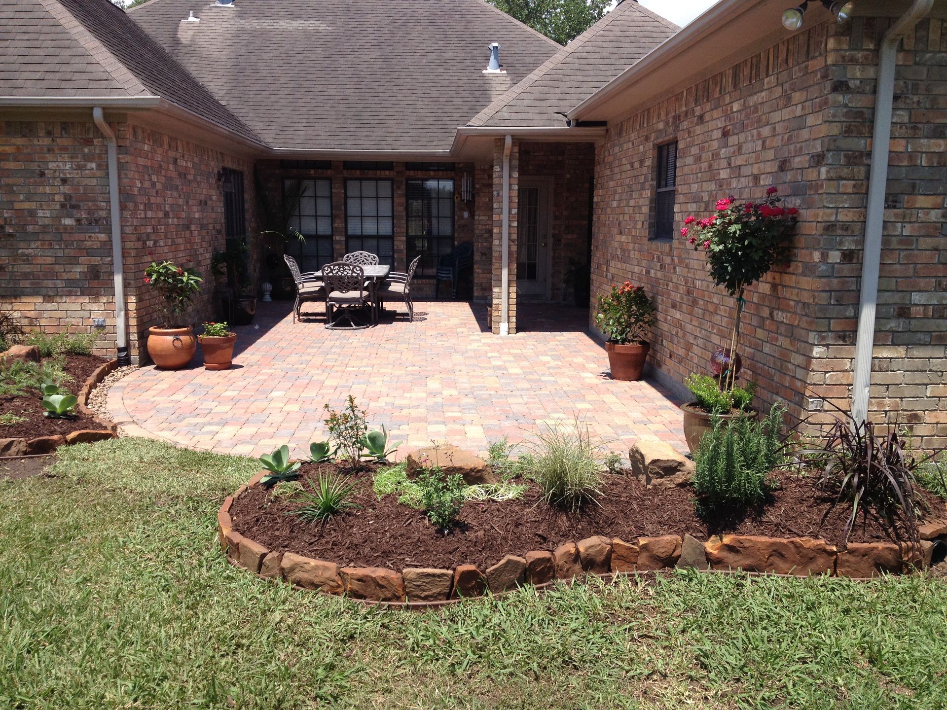 Brick patio courtyard with potted plants, flower beds, and outdoor dining set beside a house