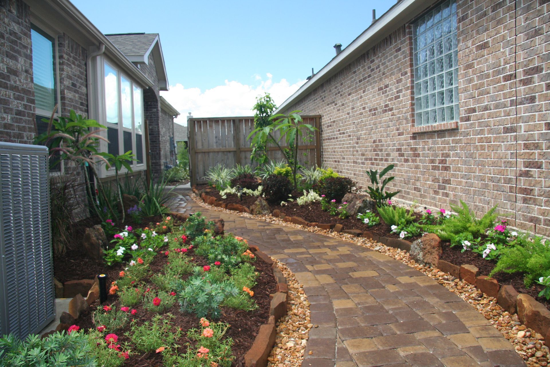 Curved brick garden path between two brick houses, bordered by flowers and greenery under a blue sky