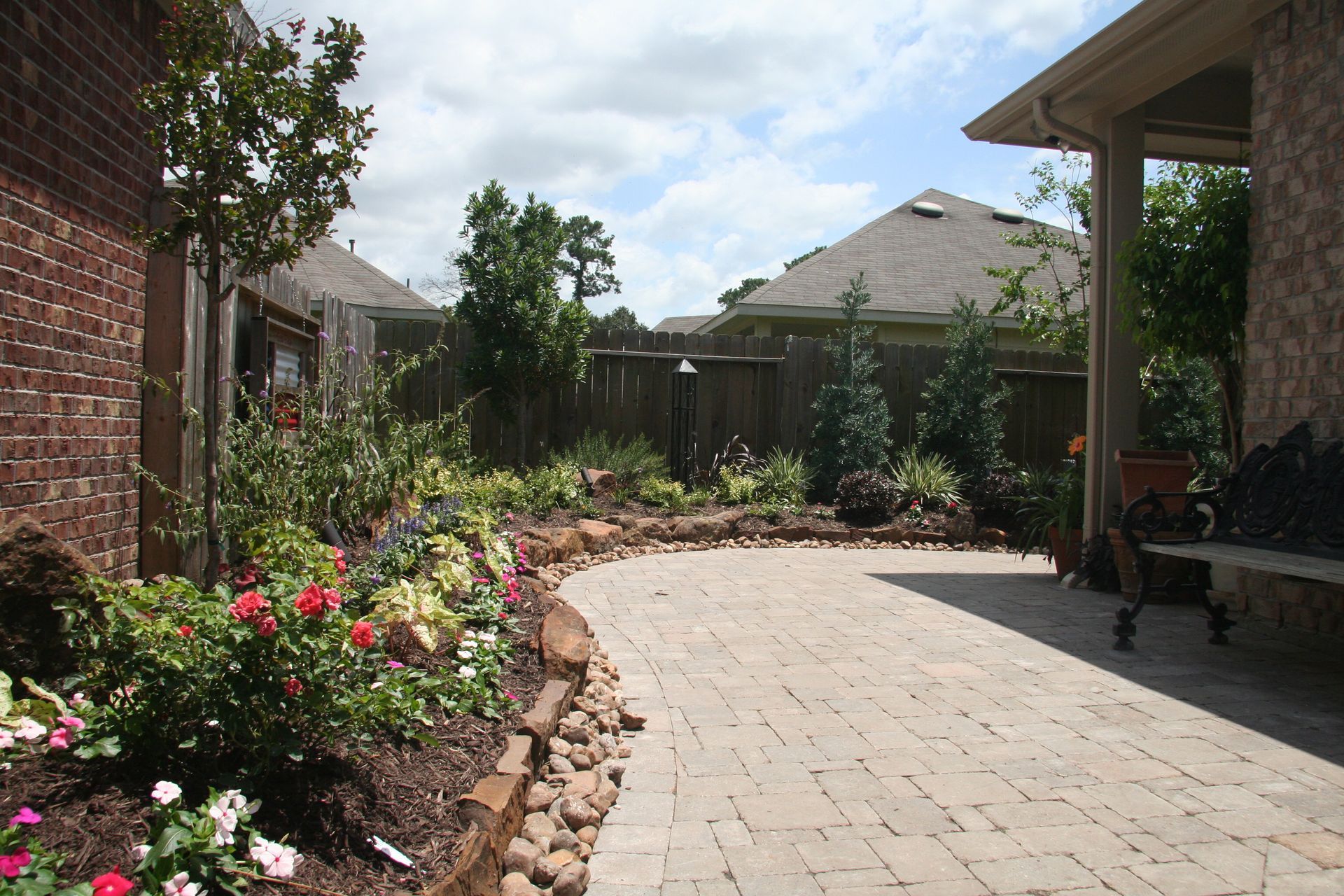 Backyard patio with stone pavers, flower beds, and a covered seating area under a sunny sky