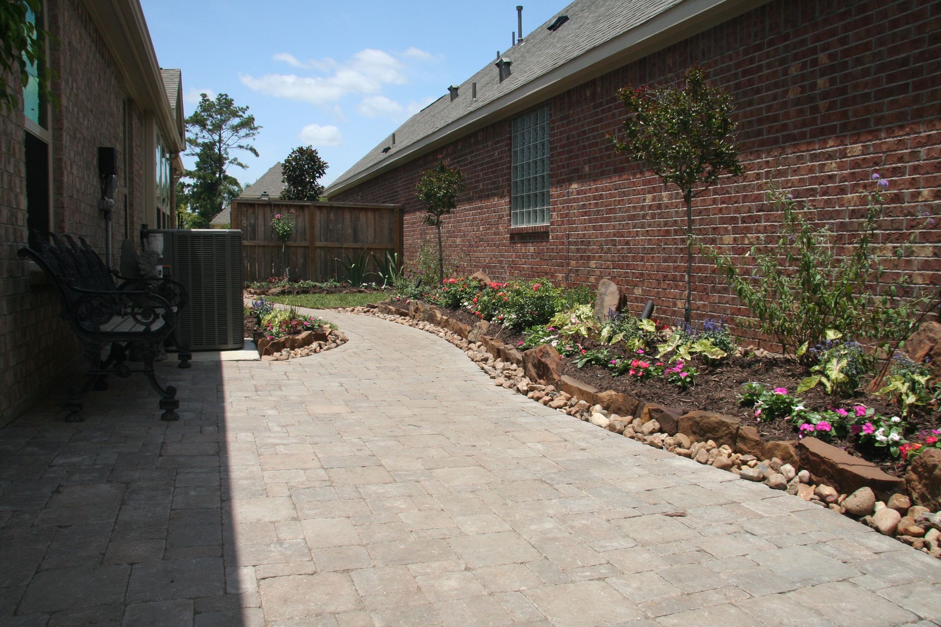 Curved brick walkway beside red brick houses, with flower beds and a wooden fence under a blue sky
