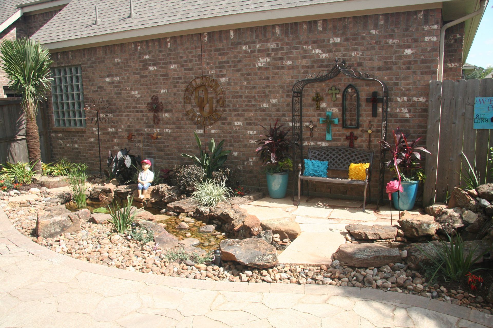 Backyard garden with stone path, flowers, small fountain, and a brick house wall