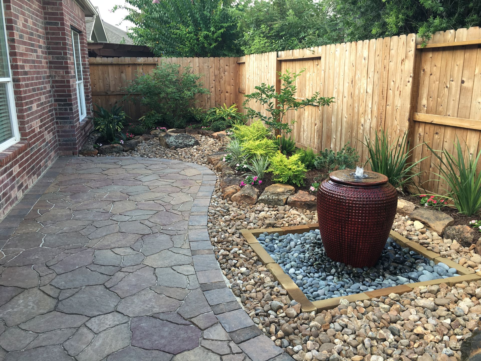 Backyard patio with stone pavers, wooden fence, plants, and a red urn fountain on a pebble bed