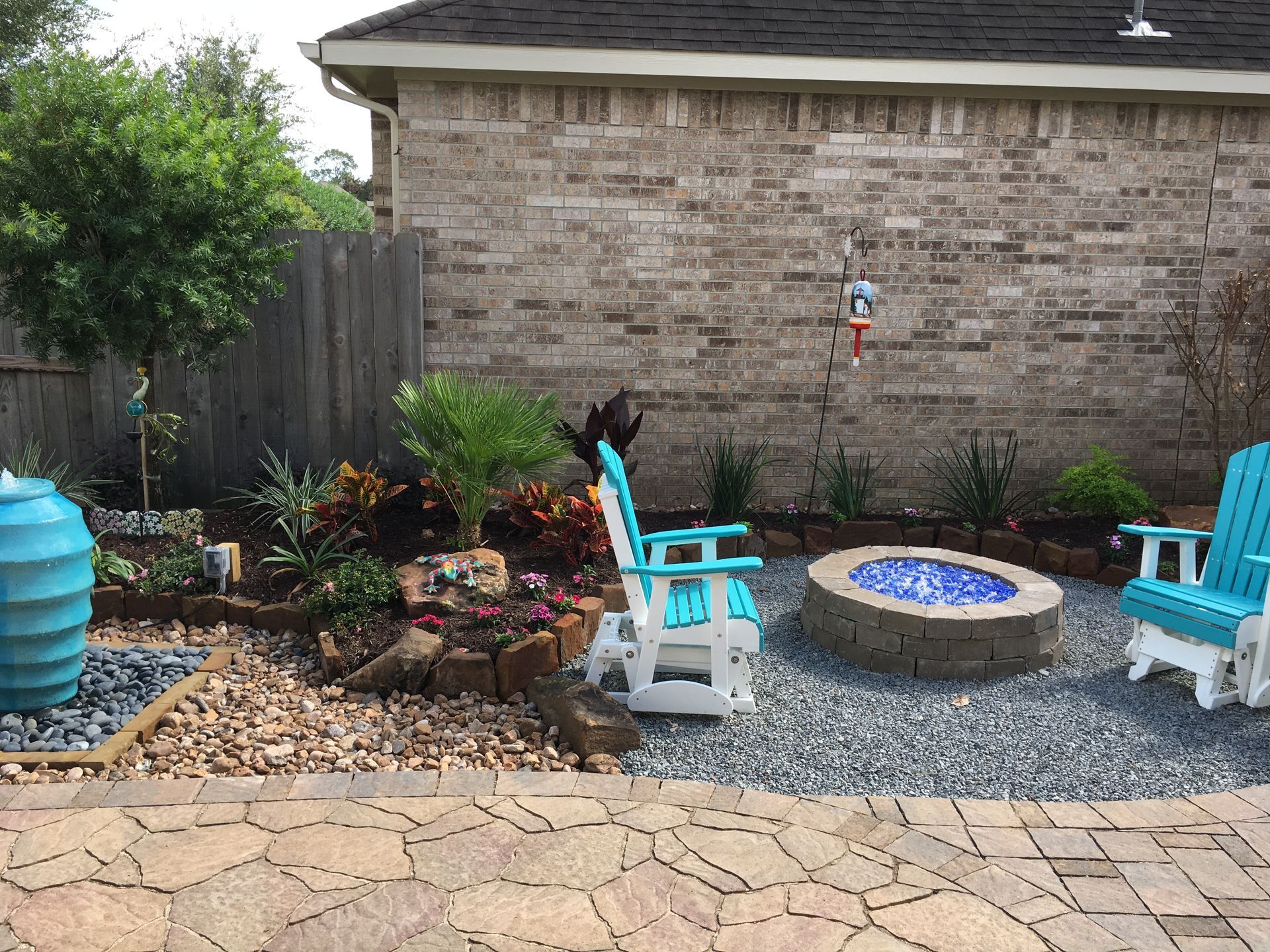 Backyard patio with stone fire pit, blue chairs, gravel, plants, and a brick wall