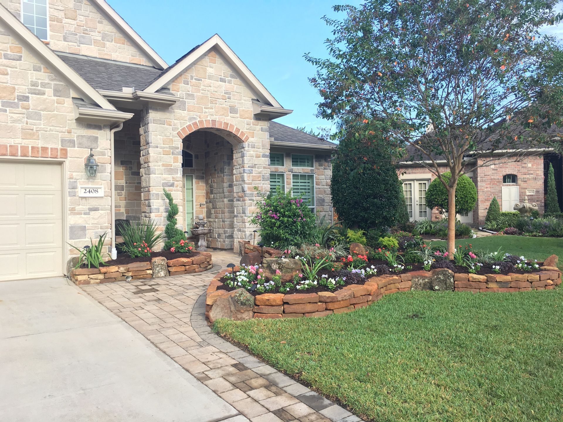 Stone house front yard with arched entry, flower beds, and a curved brick-edged lawn.