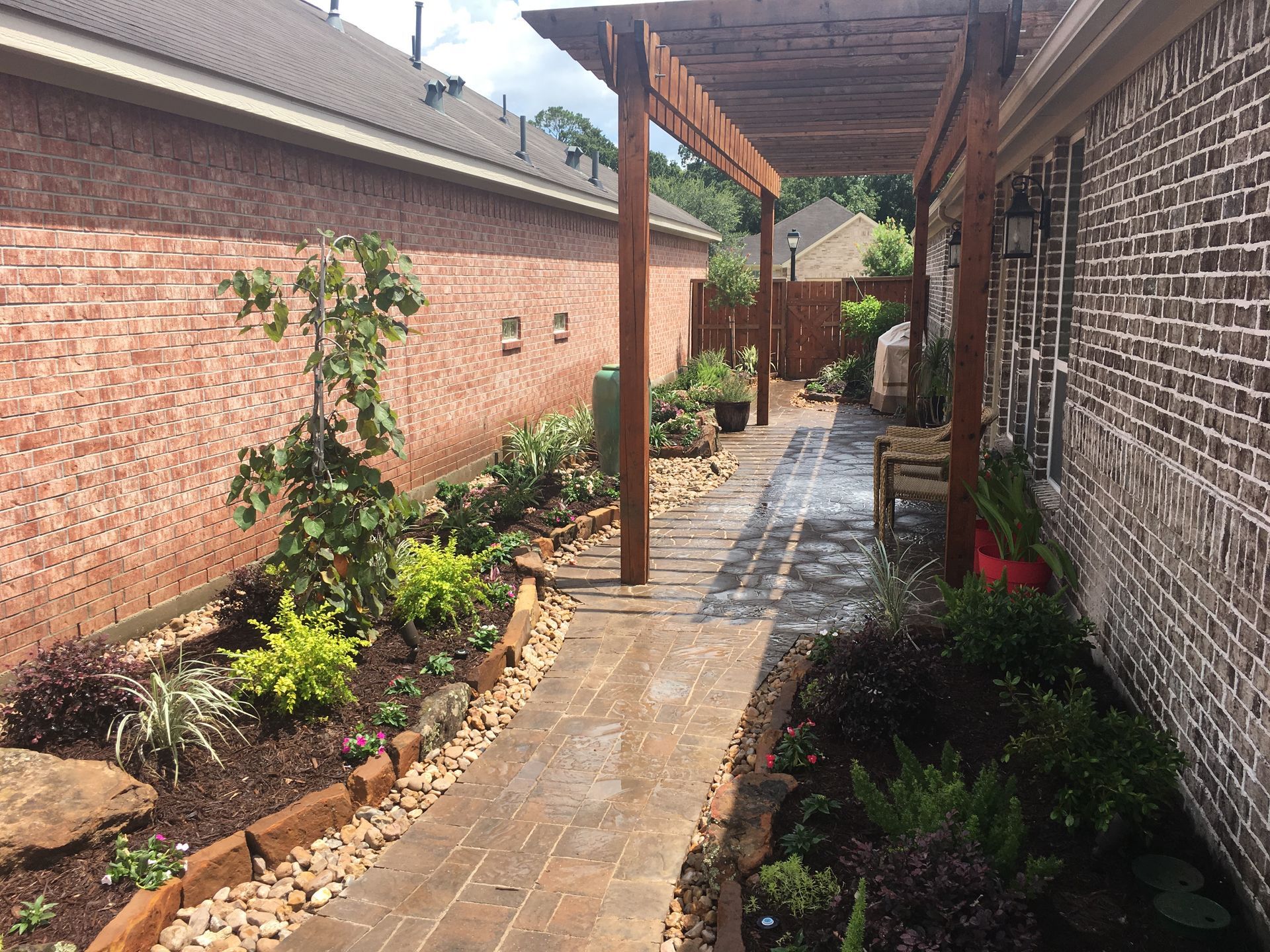 Narrow brick garden walkway with pavers, wooden pergola posts, and lush plants along a house wall