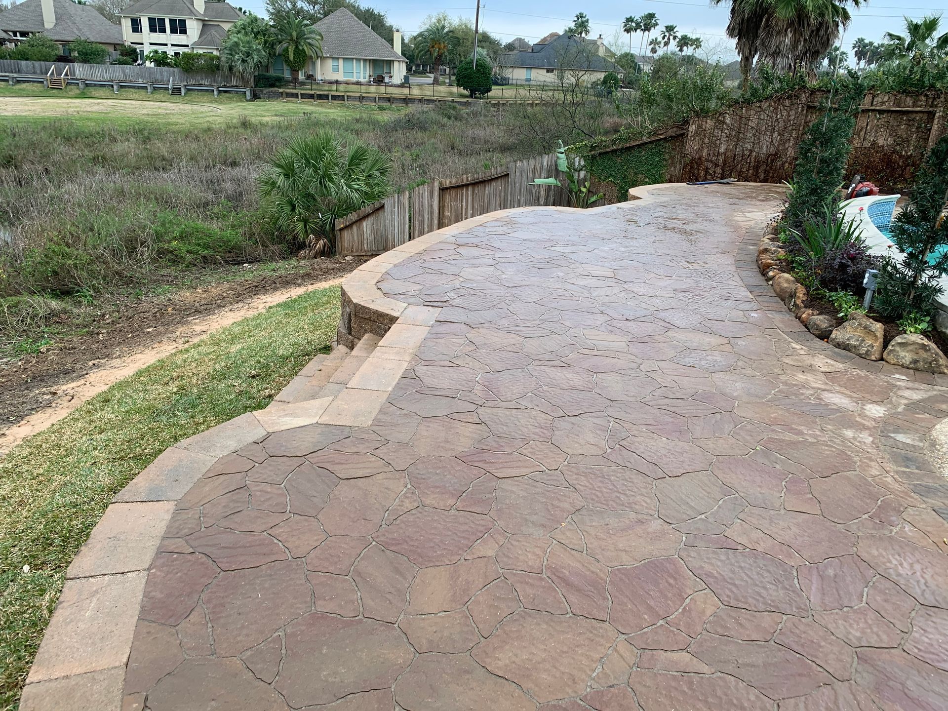 Curved stone walkway beside a grassy yard and water, with houses in the distance.