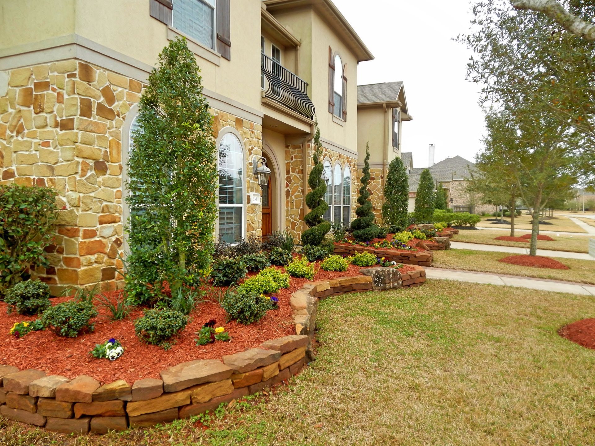 Landscaped front yard with red mulch, stone retaining wall, shrubs, and a beige house exterior