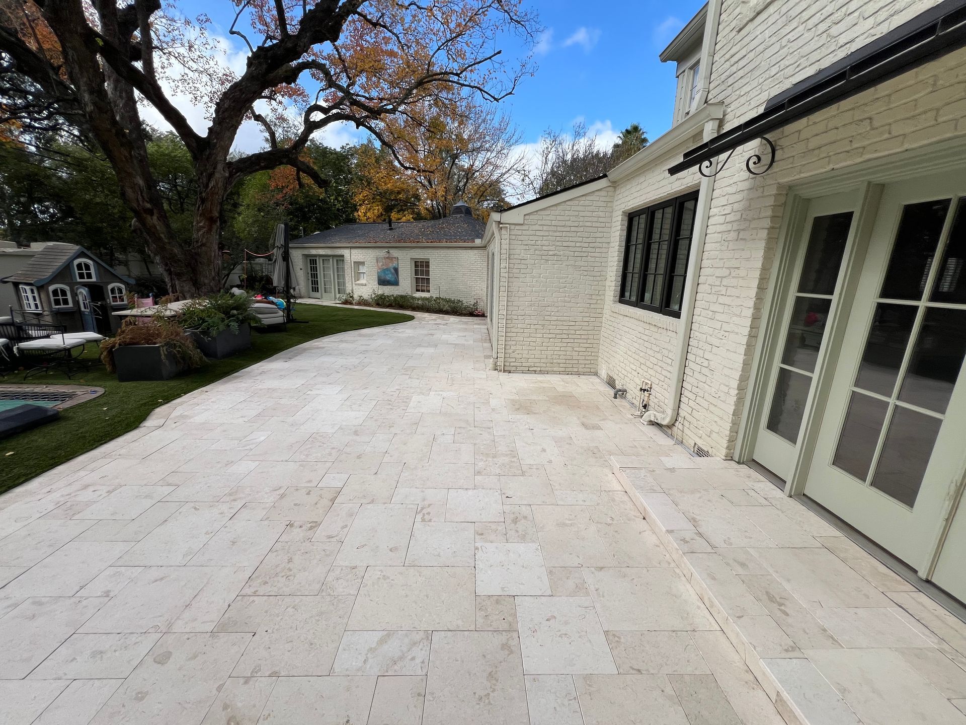 Stone patio beside a white house with glass doors, bordered by trees and a detached garage.
