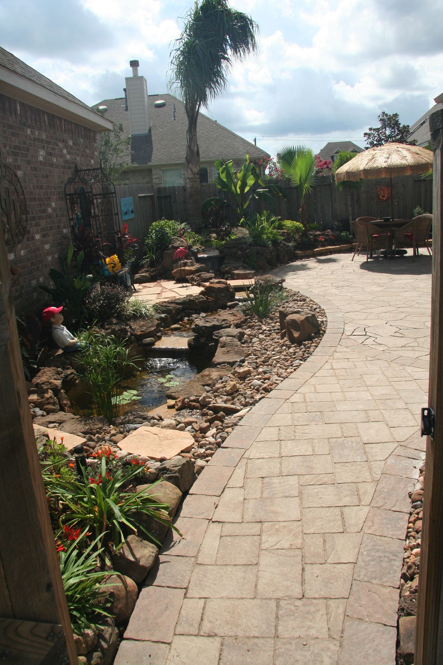 Stone path through a landscaped garden beside a house, with rocks, plants, and palm trees under a cloudy sky