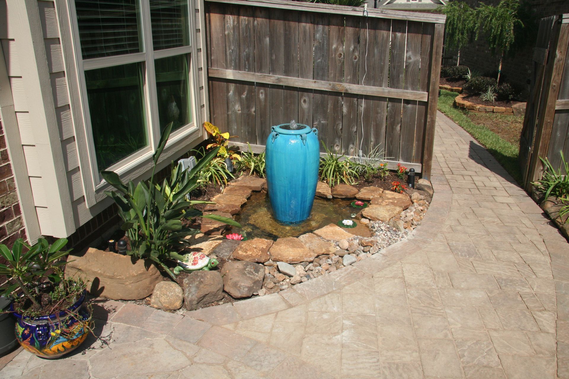 Patio with curved stone path, blue ceramic vase, potted flowers, and a wooden fence beside a house