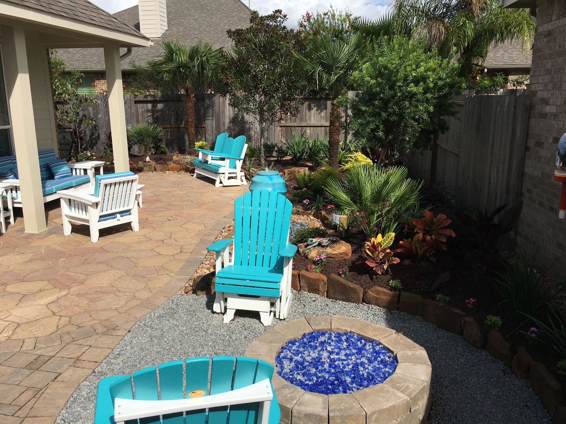 Backyard patio with turquoise chairs, a fire pit, pavers, and lush plants along a fence