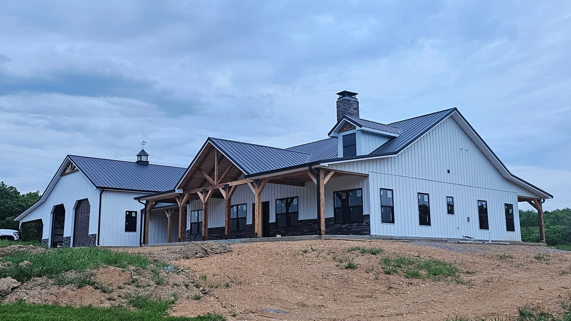 White farmhouse with dark roof and wooden porch under construction, against a cloudy sky