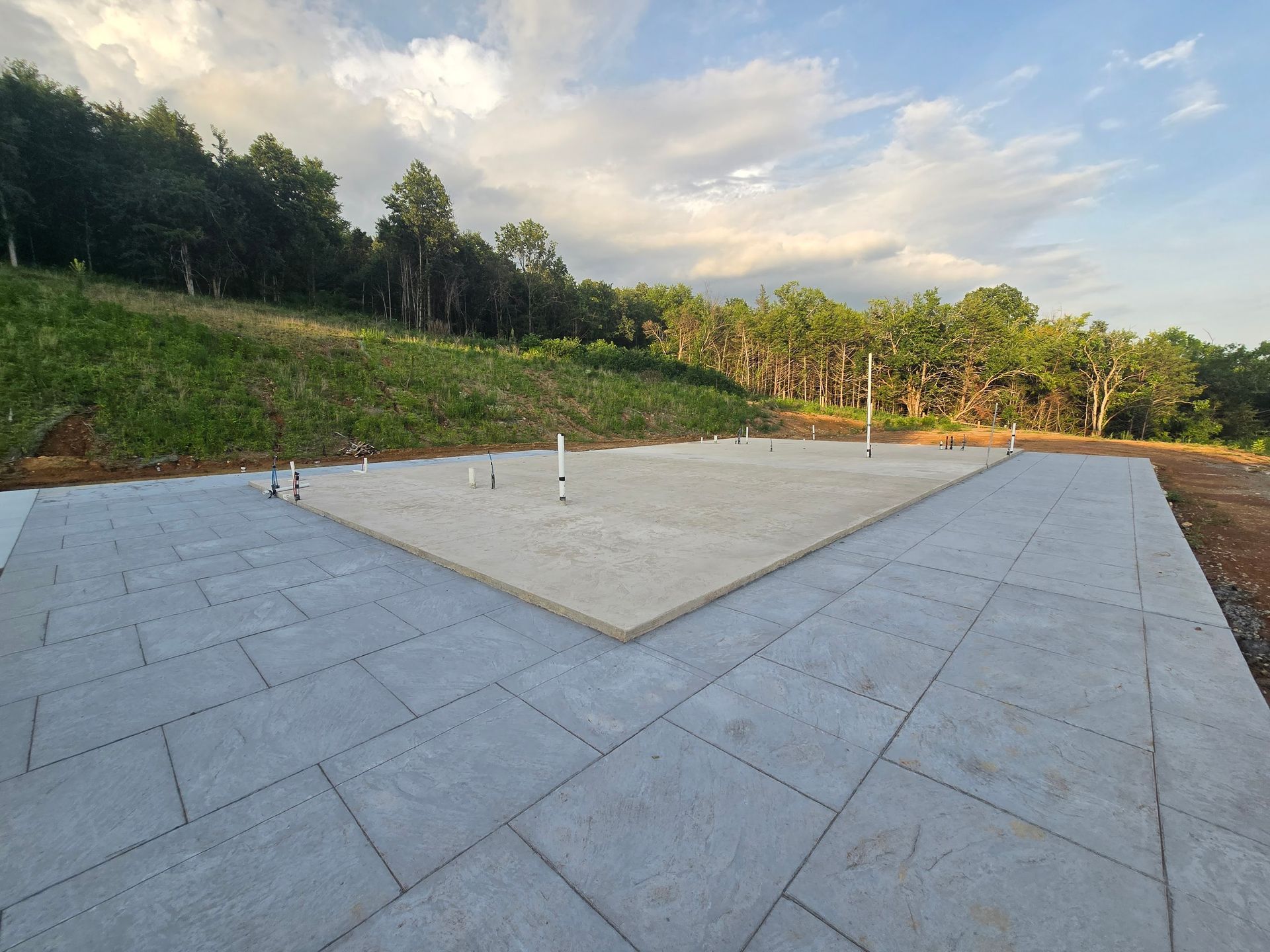 Concrete foundation with plumbing pipes surrounded by stone paving. Trees and grassy hill in the background