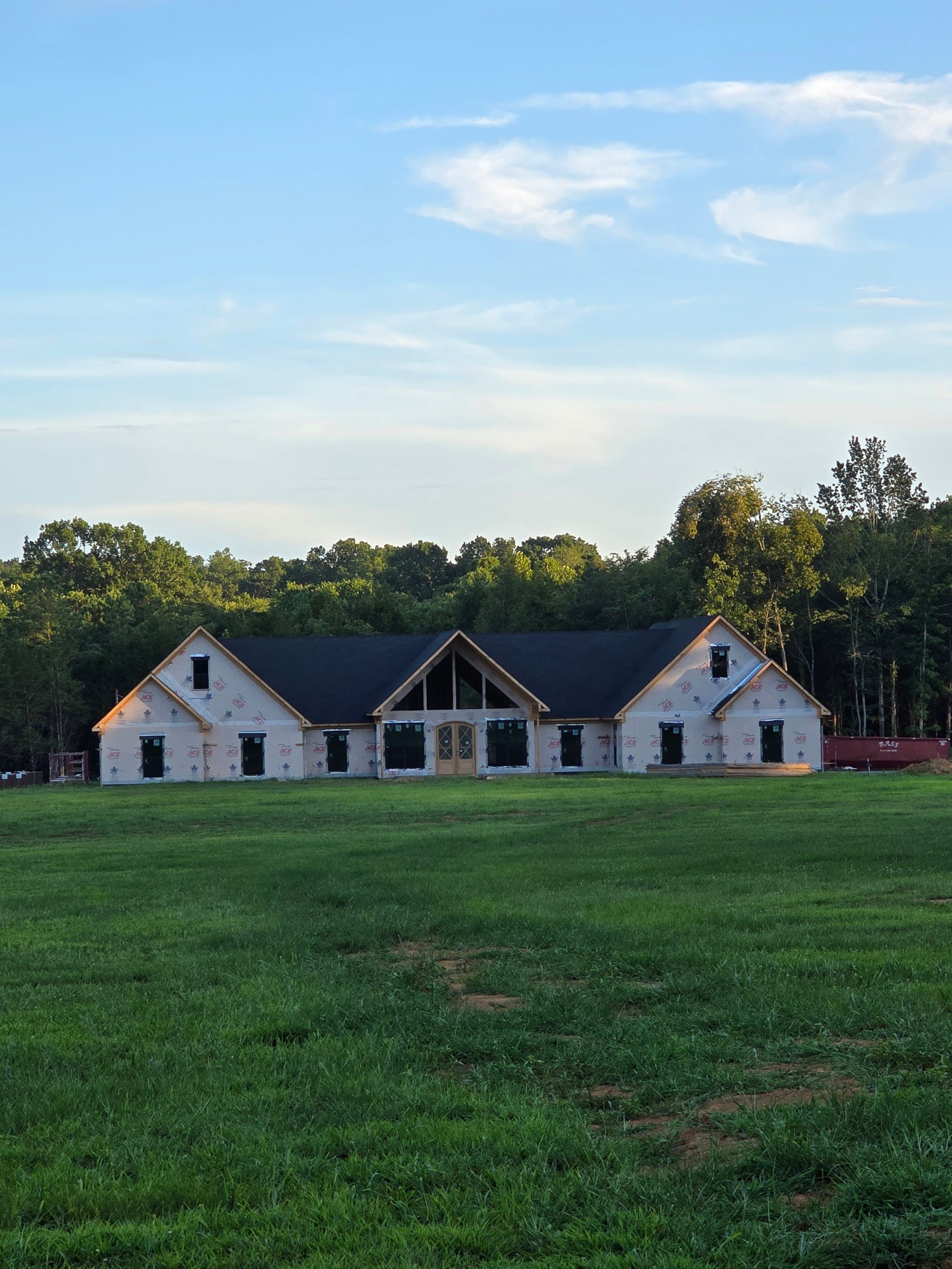 House under construction in a field, with a black roof and surrounded by trees under a blue sky