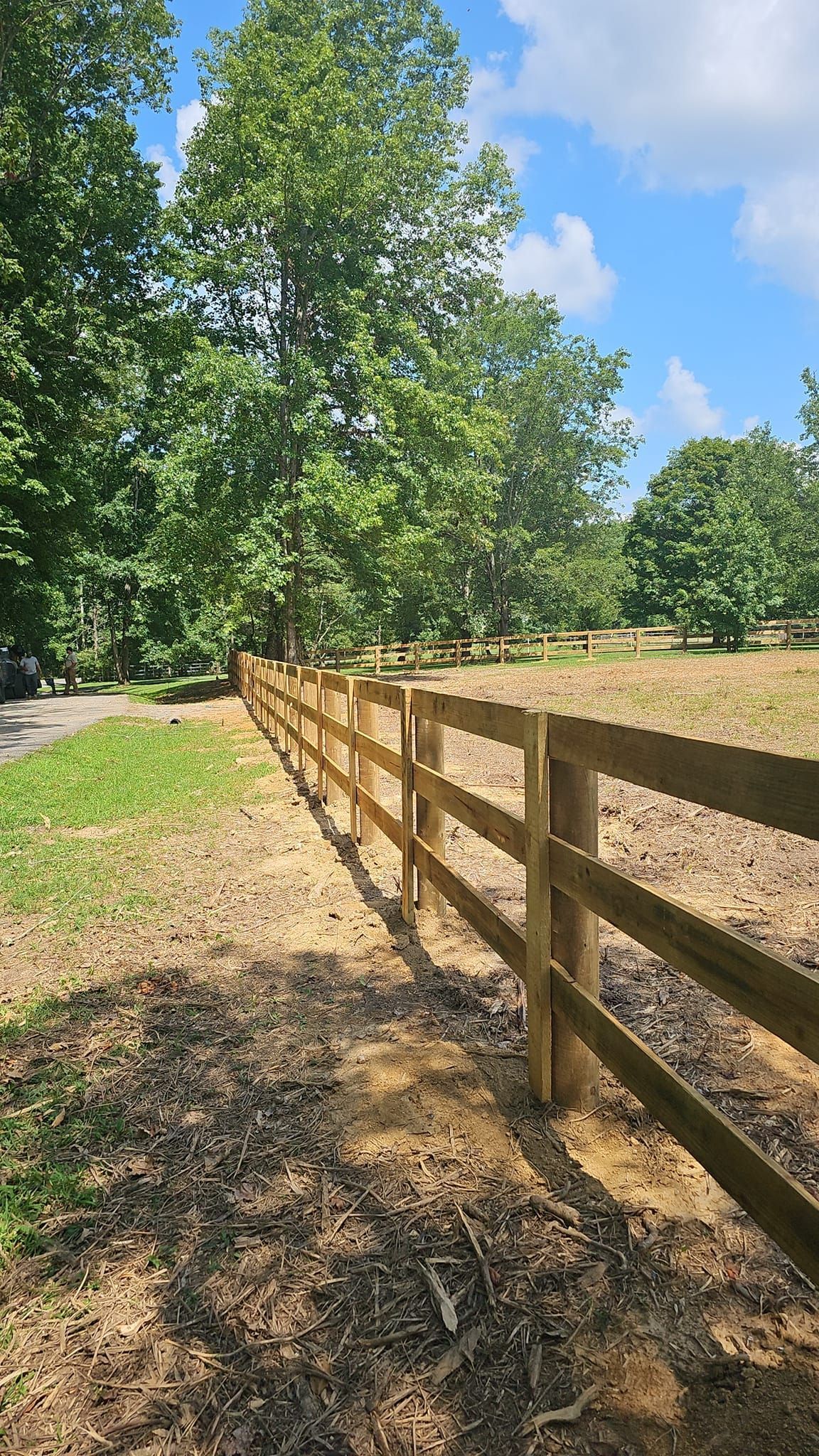 A wooden fence along a dirt road in a field with trees in the background.