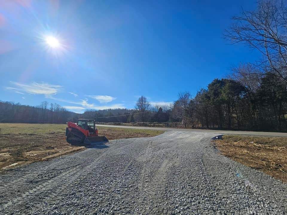 A gravel driveway leads to a road, with a small excavator on the side under a sunny, blue sky