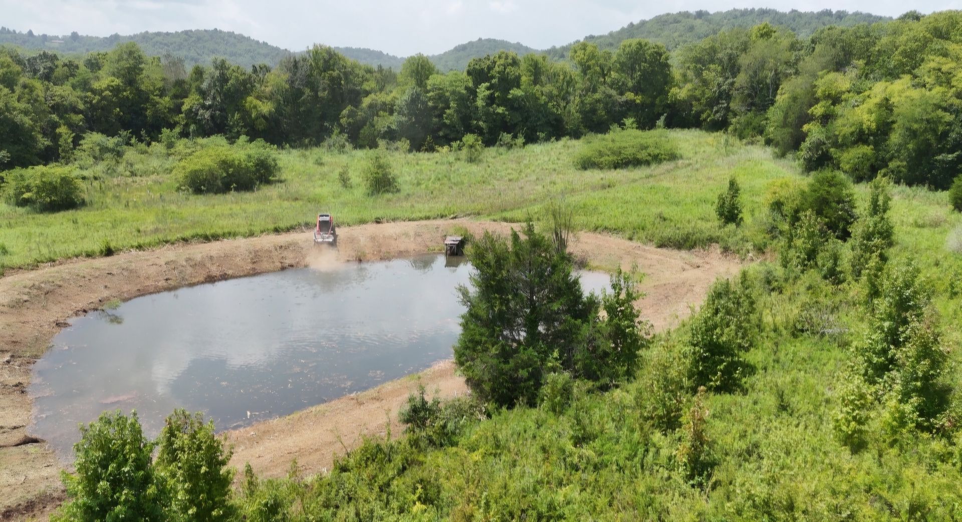 An aerial view of a small pond in the middle of a lush green field surrounded by trees.