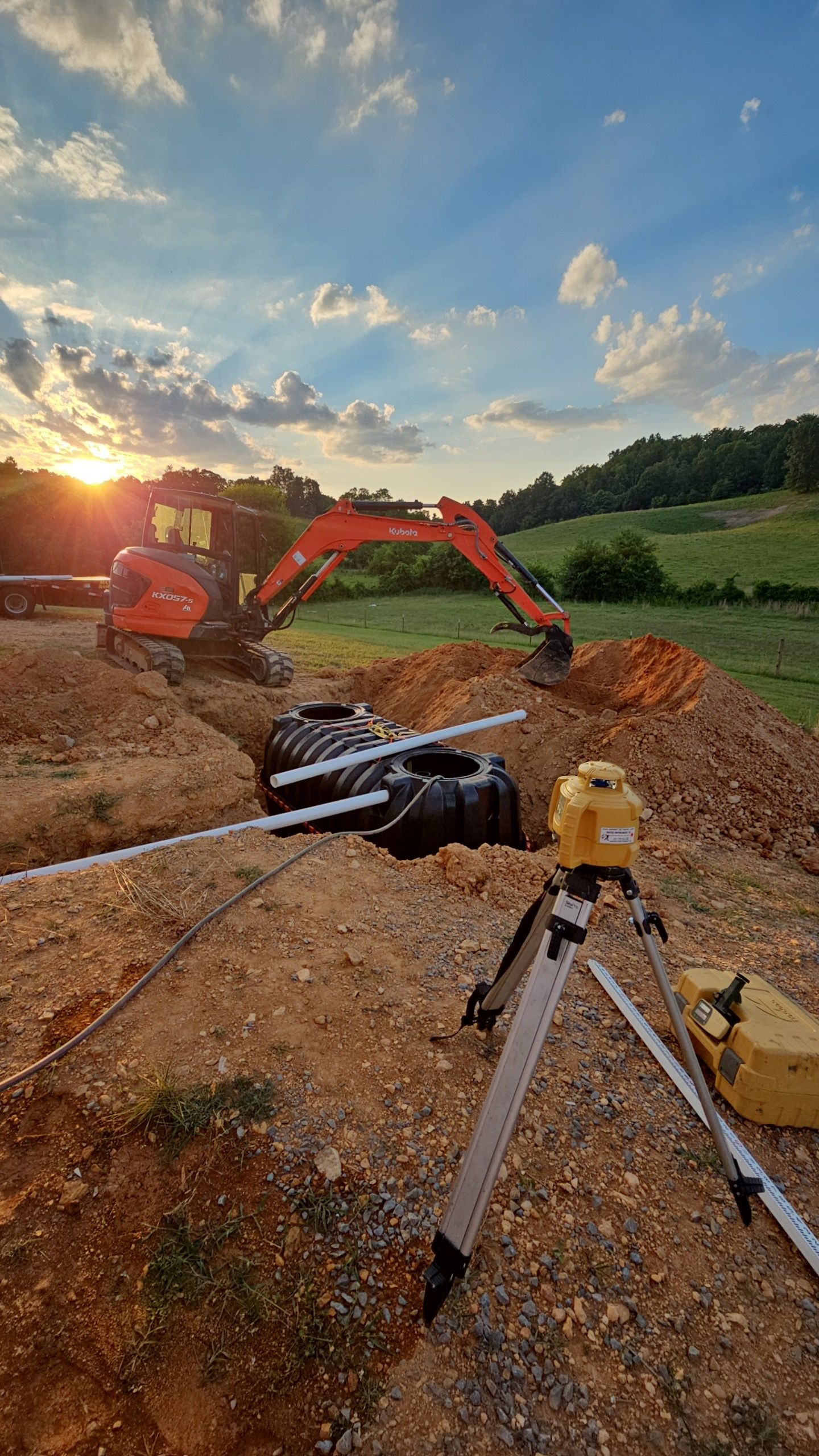 A construction site with an excavator and a tripod in the foreground and a sunset in the background.