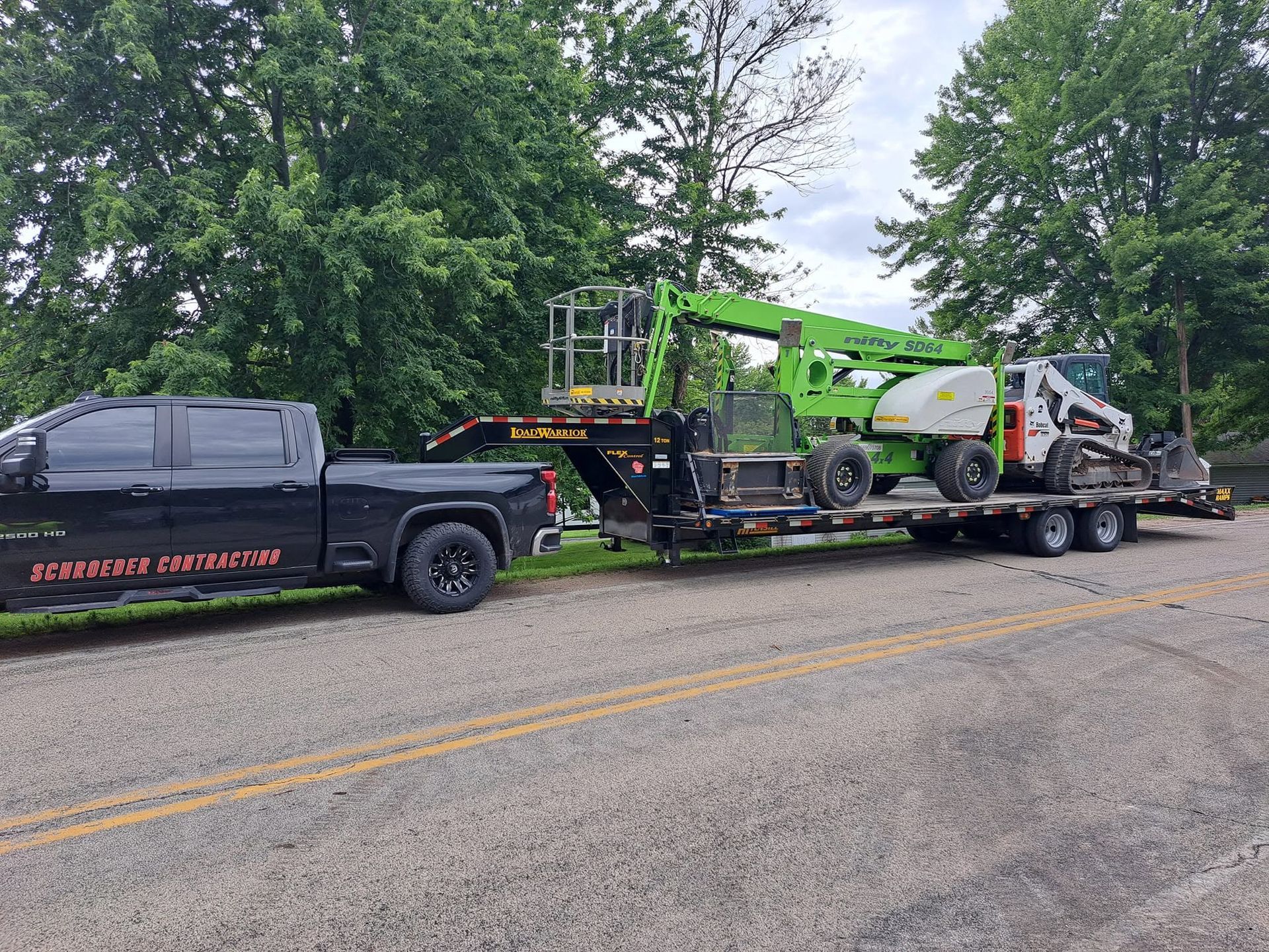 Black pickup truck towing a flatbed trailer with a green construction machine on a rural road