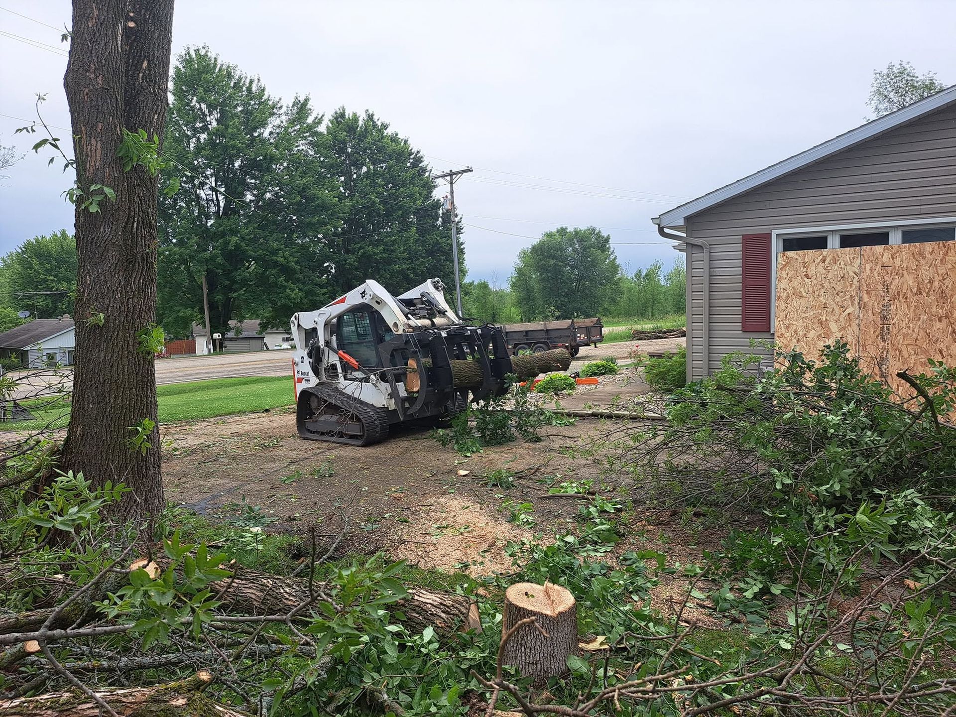 Bobcat skid steer clearing storm debris beside a house in a damaged yard with fallen branches.