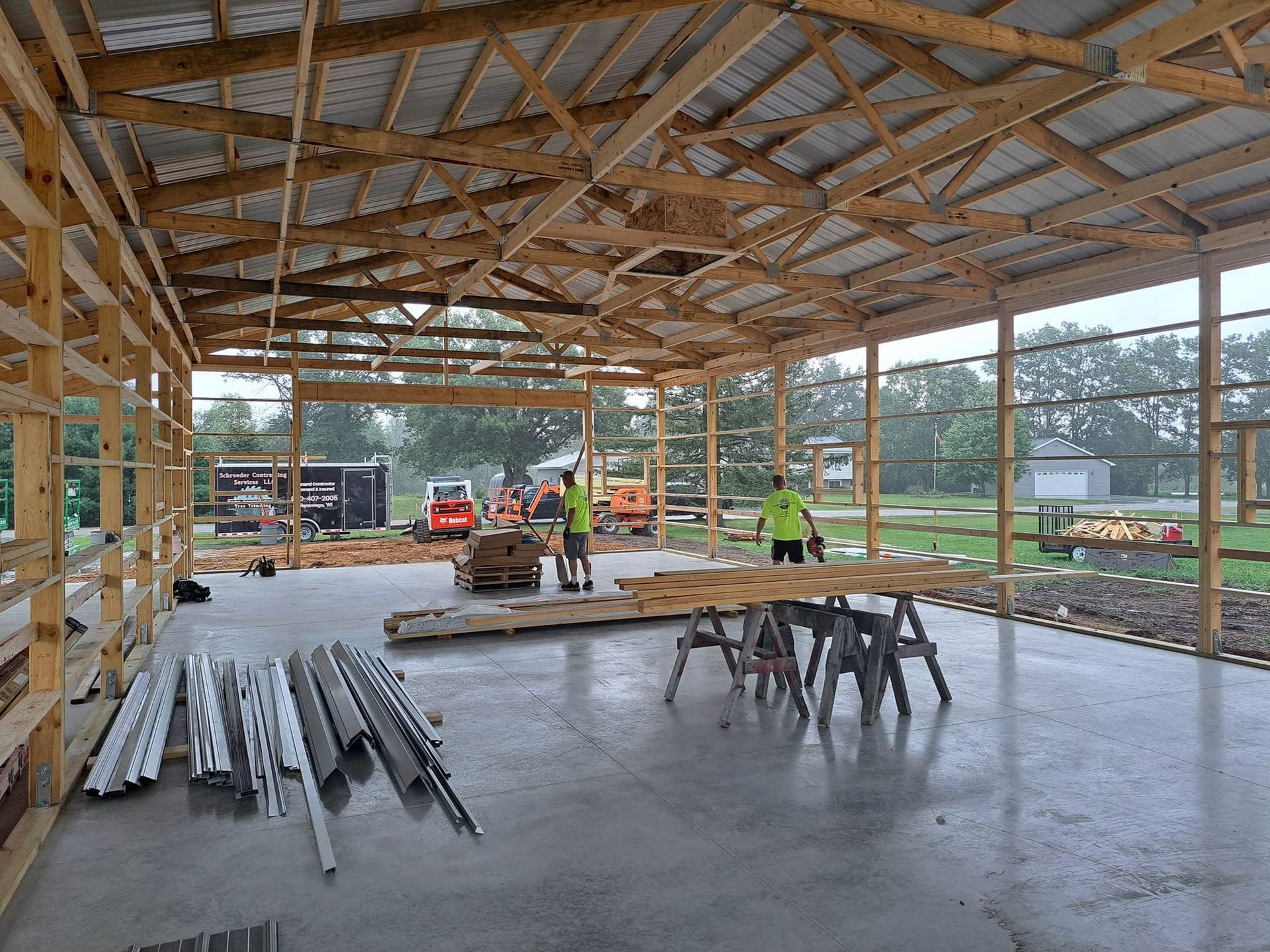 Workers framing a large open-sided wooden building with a concrete floor and stacked lumber inside.