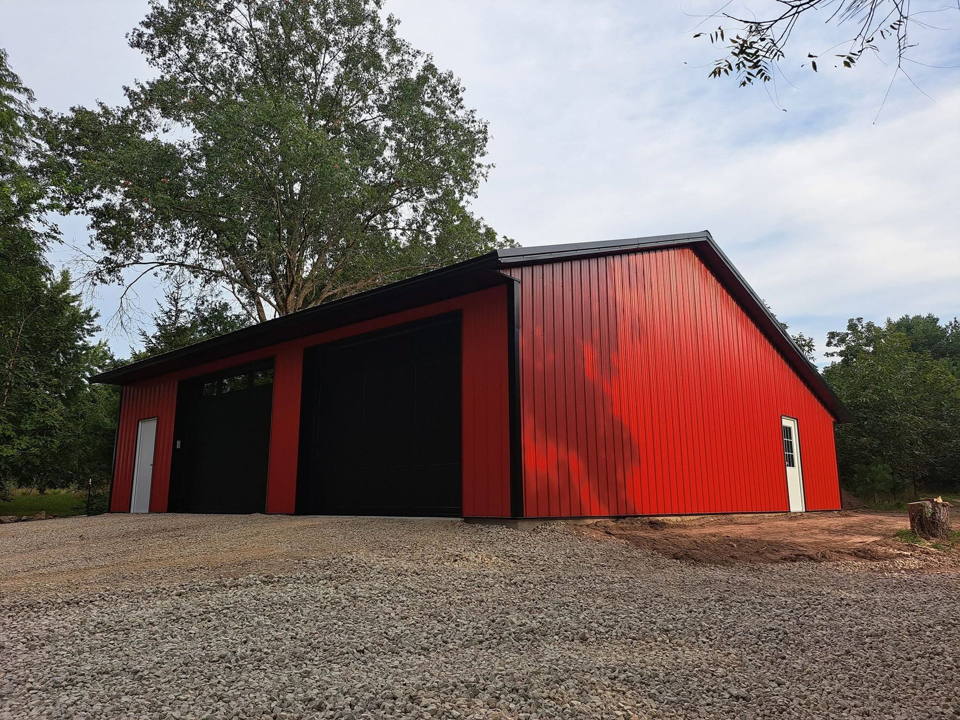 Red metal barn with black doors on a gravel lot, surrounded by trees under a cloudy sky
