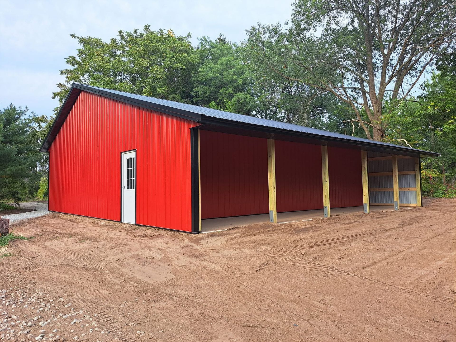 Red metal barn with open side bays on a dirt lot, surrounded by trees.