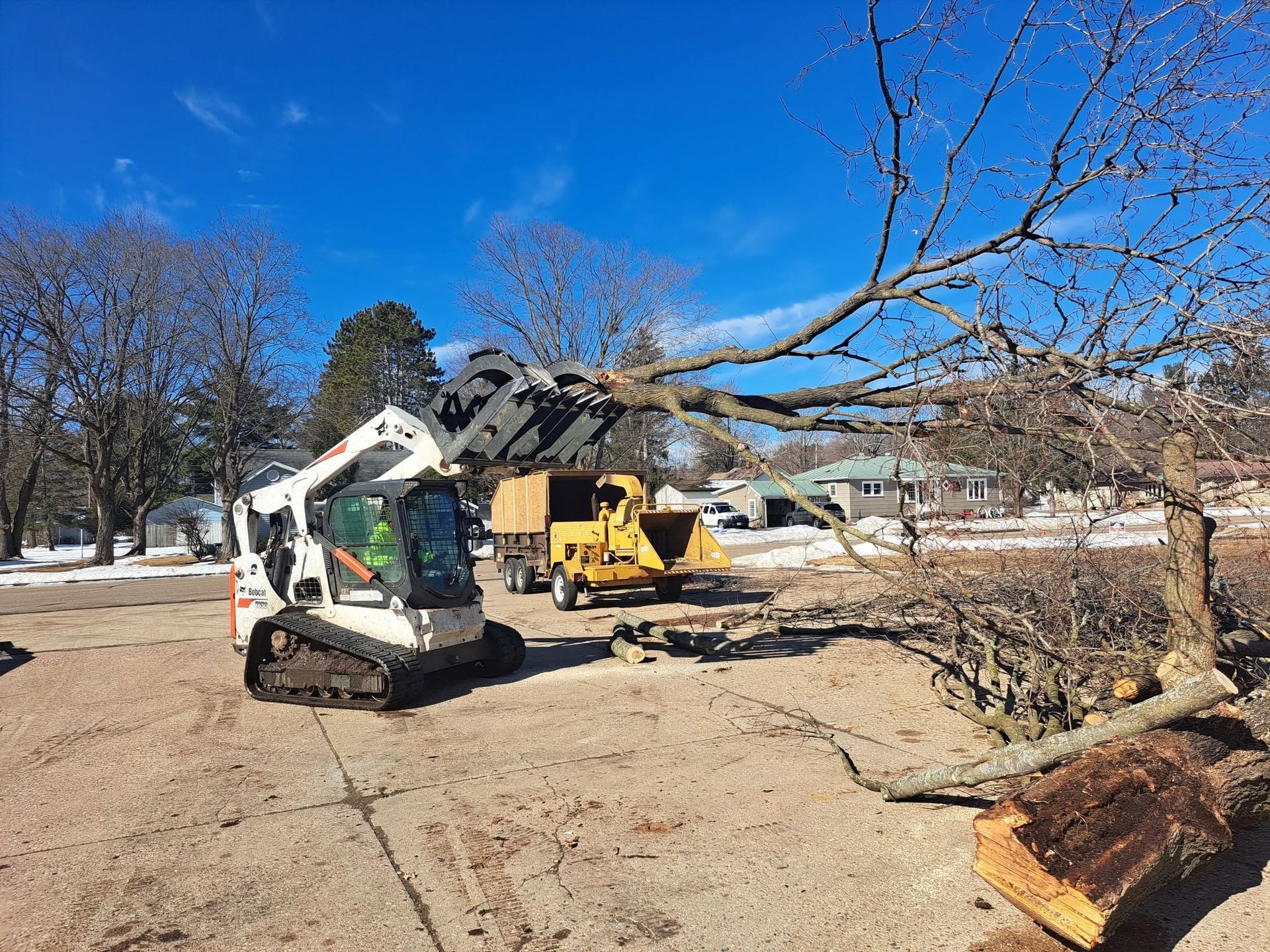 Tree-removal crews using a skid steer and crane to clear a fallen tree in a snowy yard