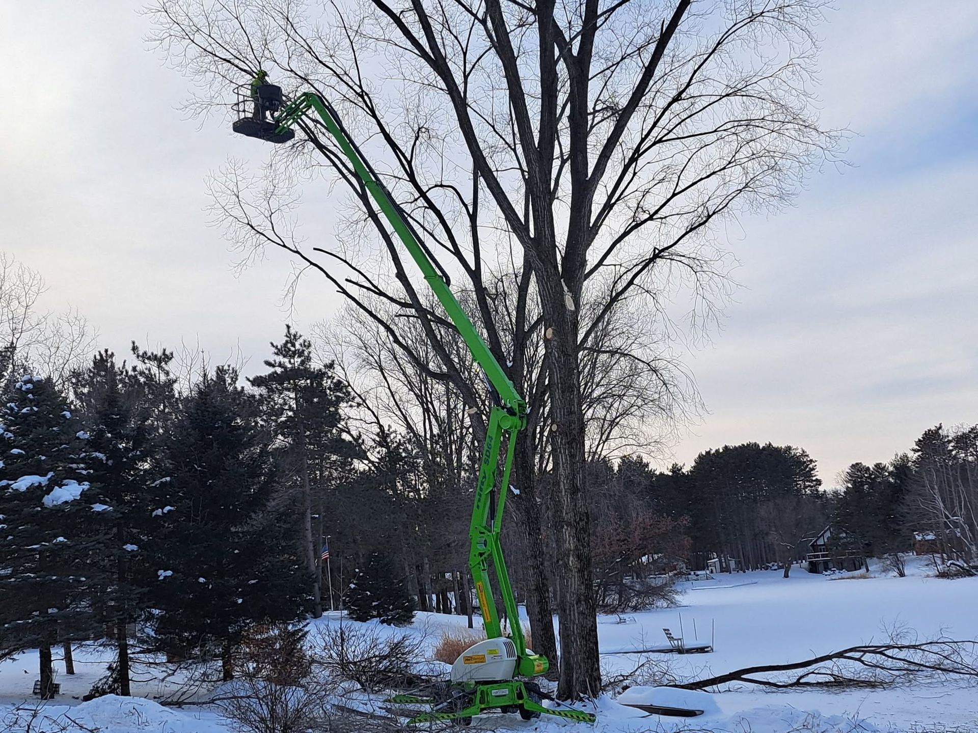 Green cherry picker lifting a worker beside a leafless tree in a snowy yard