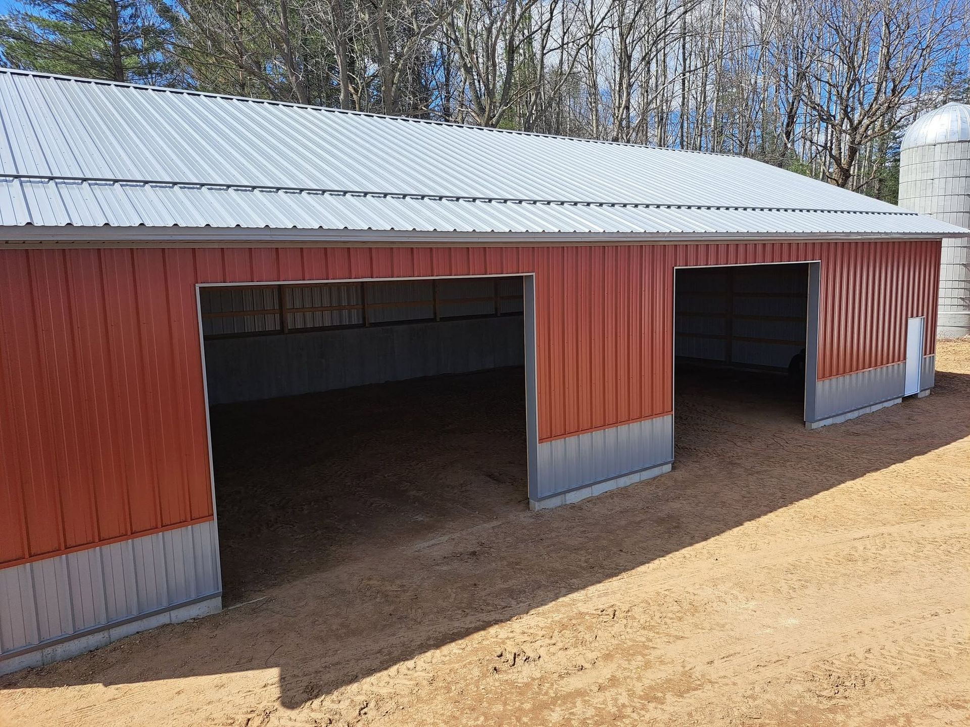 Red barn with two open garage bays and a white metal roof on a dirt lot