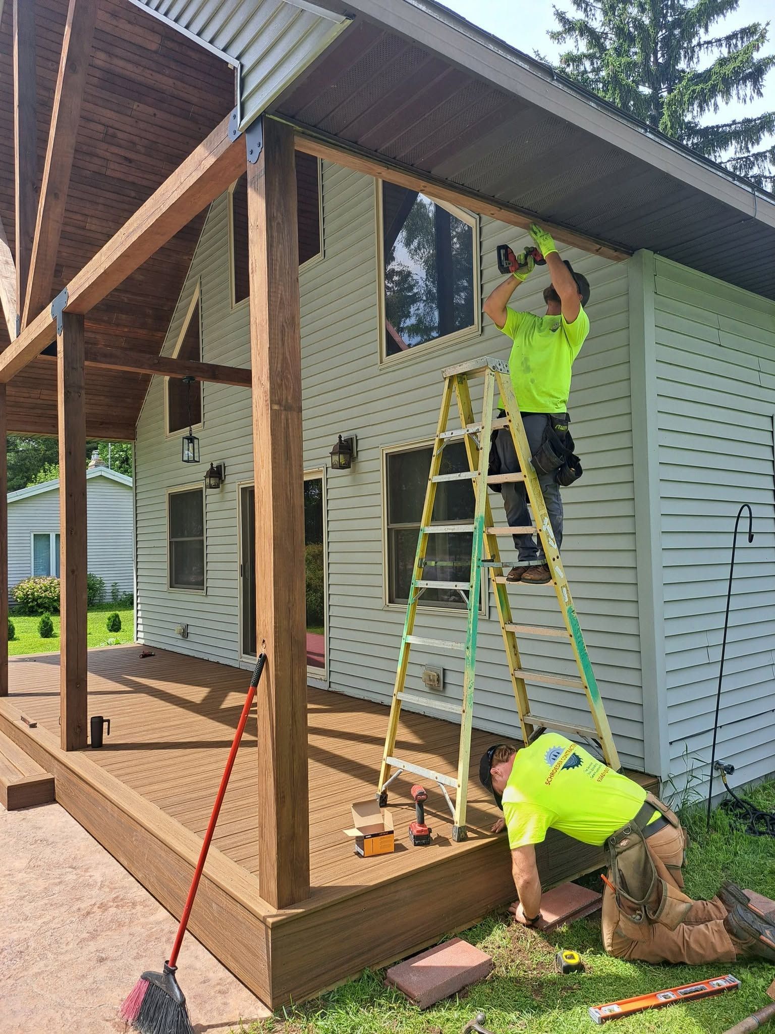 Workers installing siding on a house porch, one on a ladder and one kneeling by the deck.