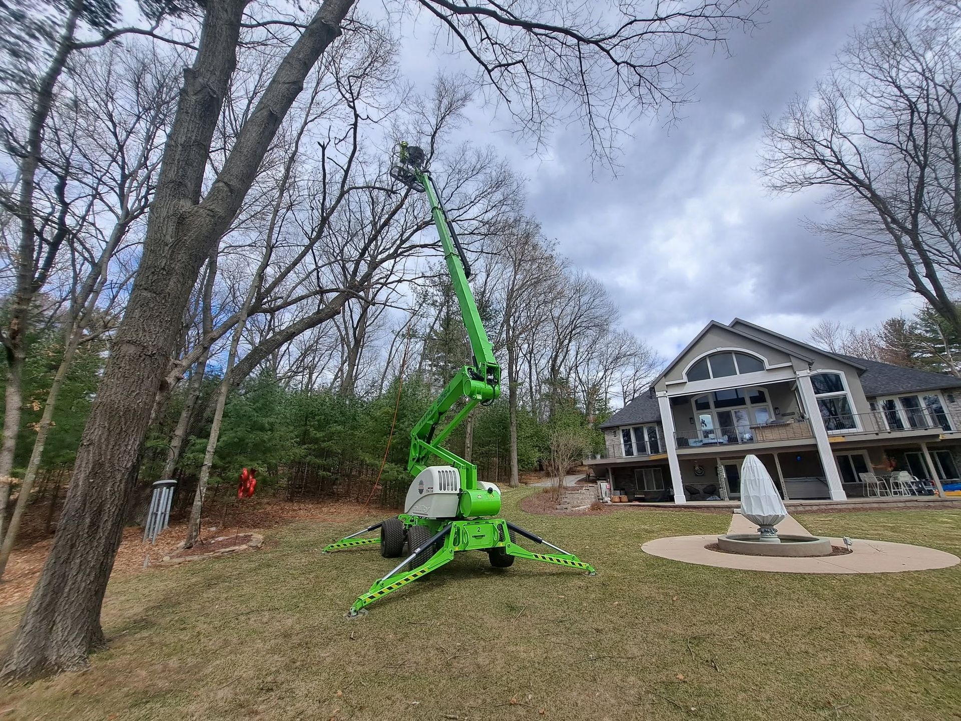 Bright green crane lifting a tree in a wooded yard beside a house with a fountain and deck