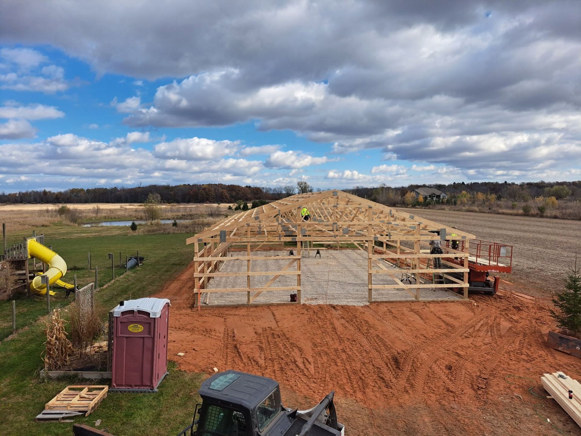 Wood-framed house under construction on a red dirt lot beneath a cloudy sky