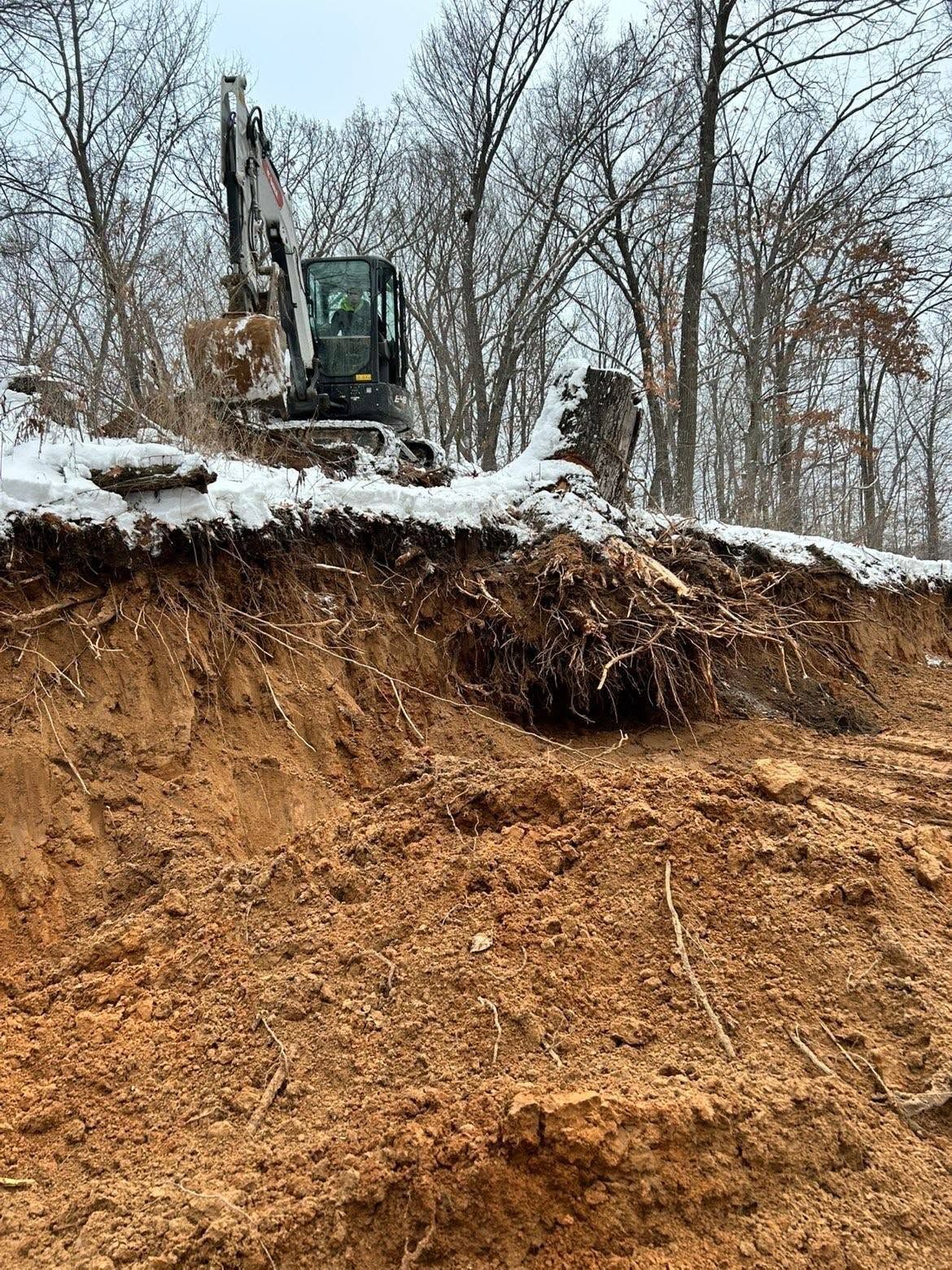 Excavator perched on a snowy cliff above a steep, eroded dirt embankment in a leafless winter forest