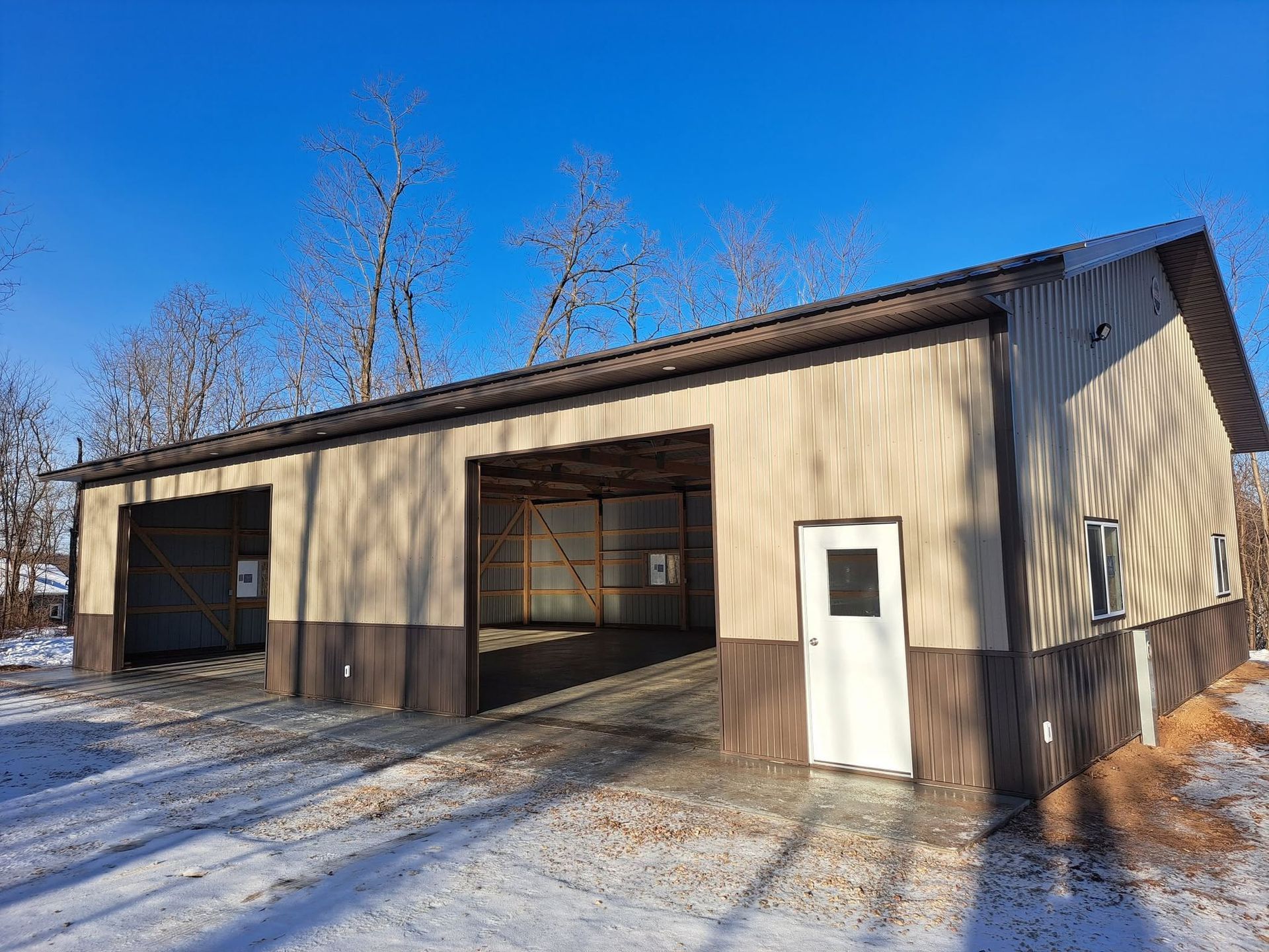 Snowy rural garage building with open bays and a white side door under a clear blue sky