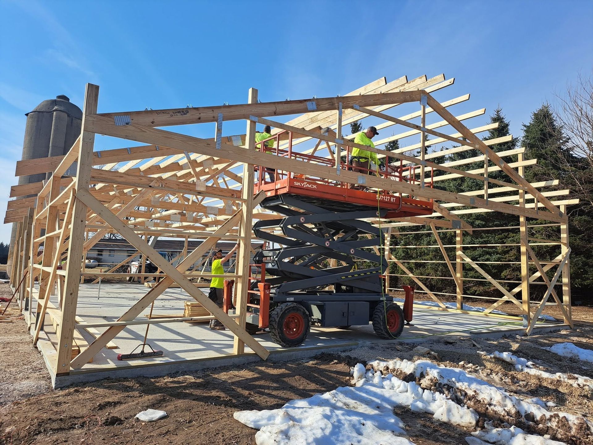 Wood-framed building under construction with workers on a lift, outdoors in snow-covered ground.