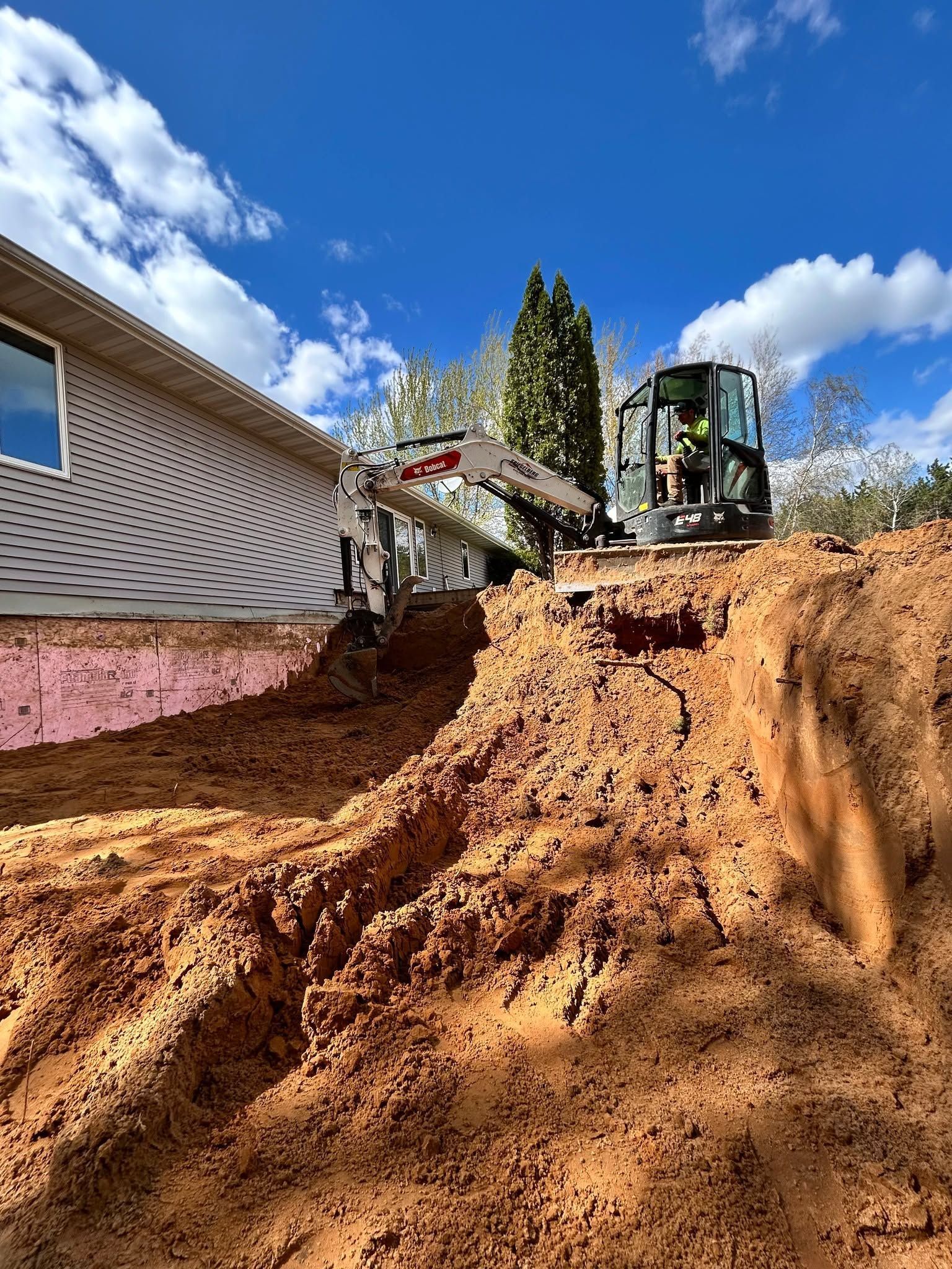 Construction excavator beside a house, digging into a steep orange dirt trench under a blue sky.