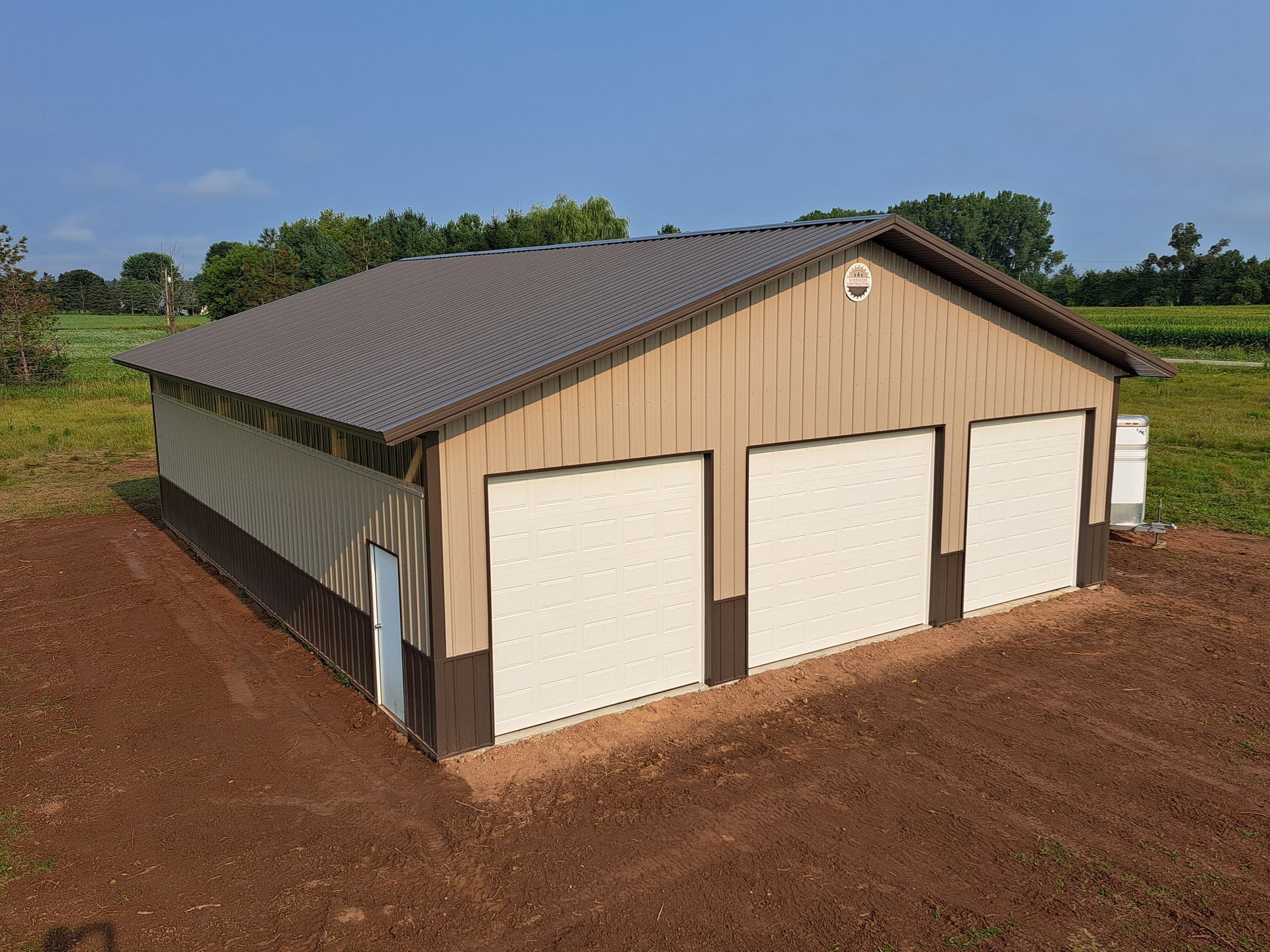 Tan metal farm building with white garage doors on a dirt lot under a clear sky