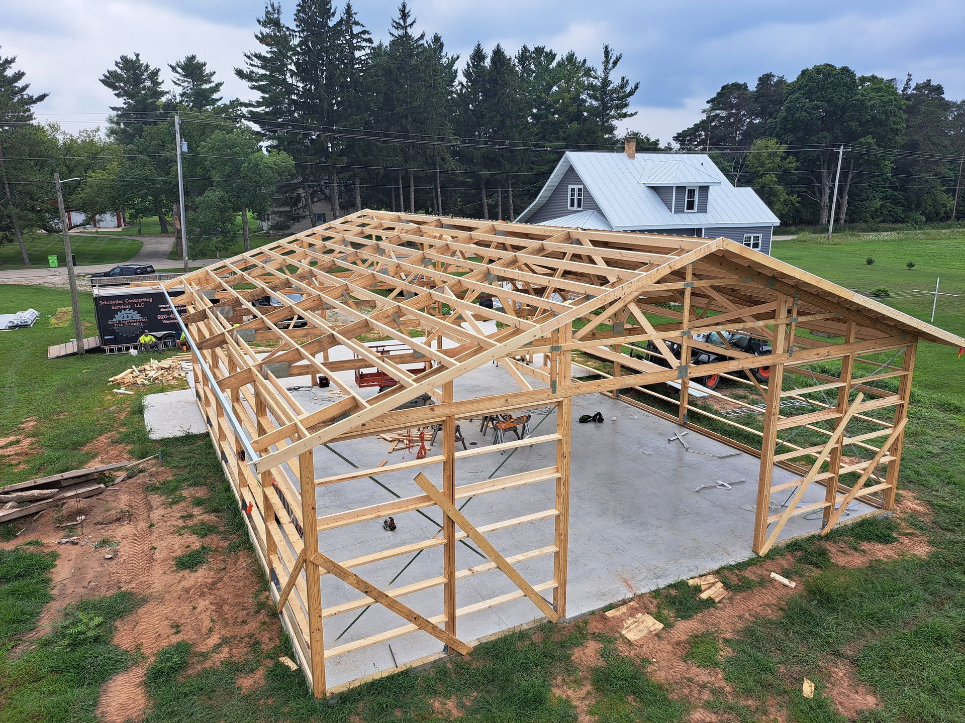 Wooden barn frame under construction on a concrete slab beside a white house and trees