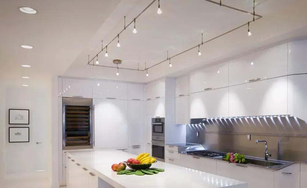 Modern white kitchen with island, fruit bowl, built-in oven, and stainless-steel counters under track lighting