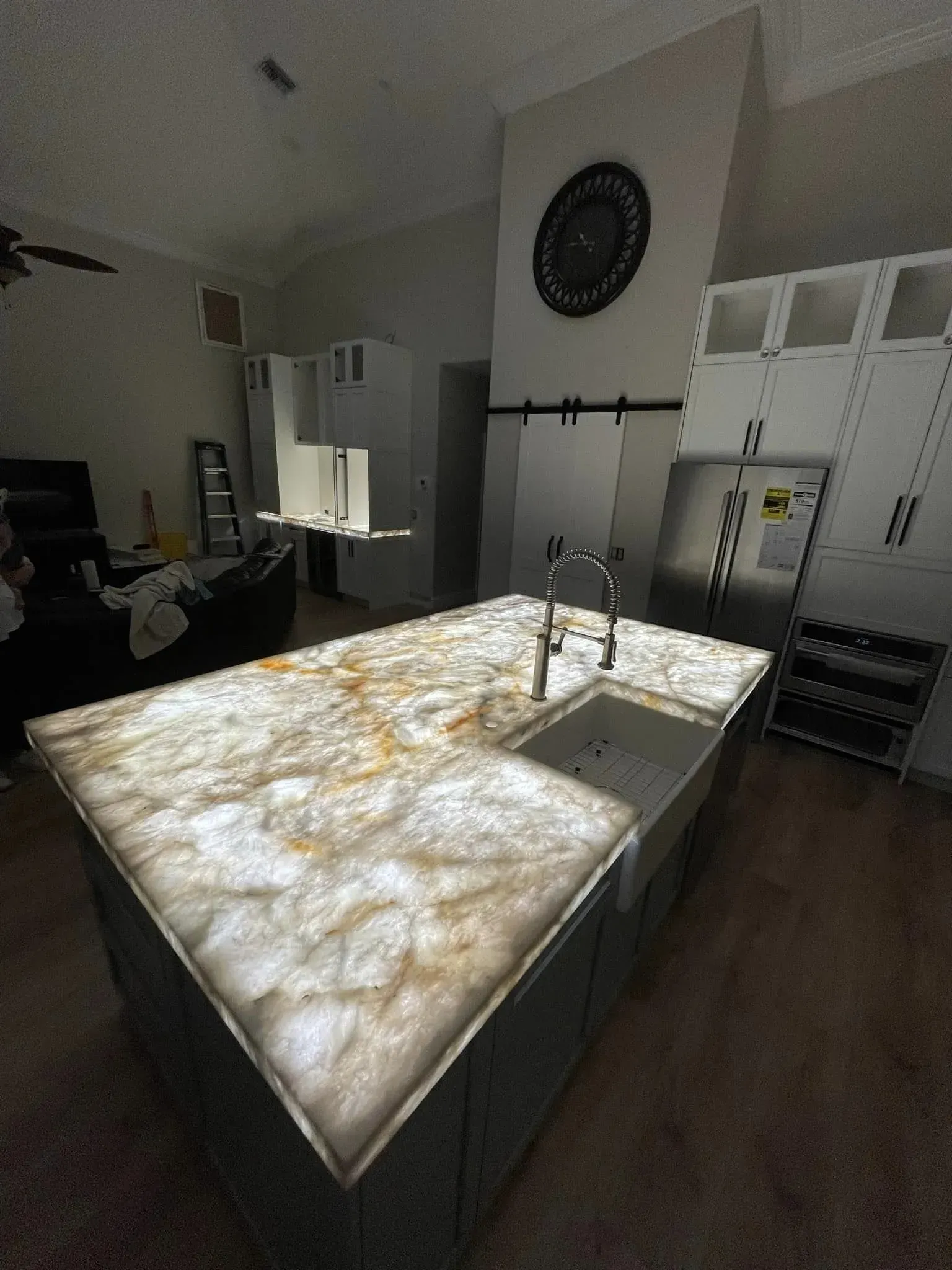 Modern kitchen with a large marble island, sink, white cabinets, and sunlight streaming through a window.