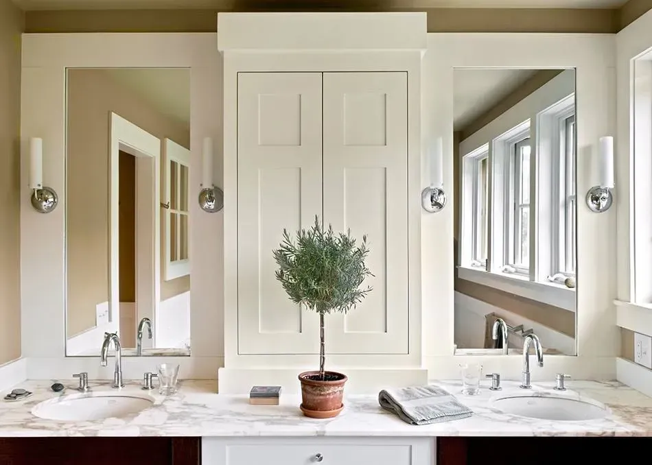 Symmetrical bathroom vanity with double sinks, mirrors, and a potted plant centered between white cabinets.