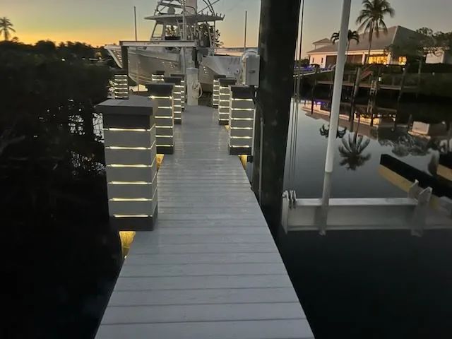 Illuminated dock walkway at dusk beside calm water and moored boats, with palm trees in the background
