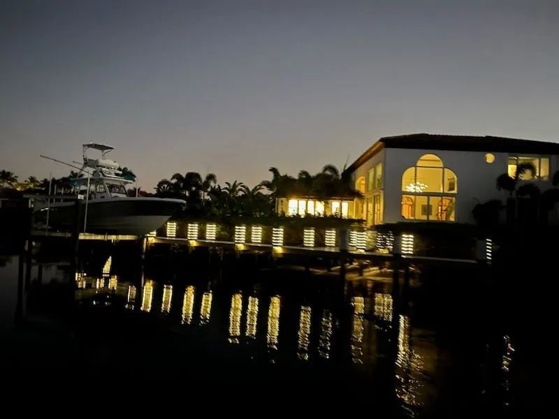 Evening waterfront resort with lit building and palm trees reflecting on calm water