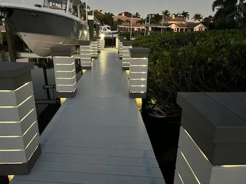 Illuminated dock walkway at dusk with steps and palm-lined waterfront homes in the background