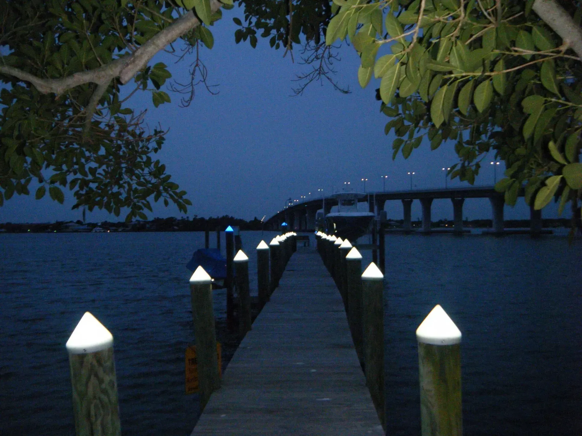 Lit wooden pier at dusk with glowing white post lights leading to a bridge over calm water