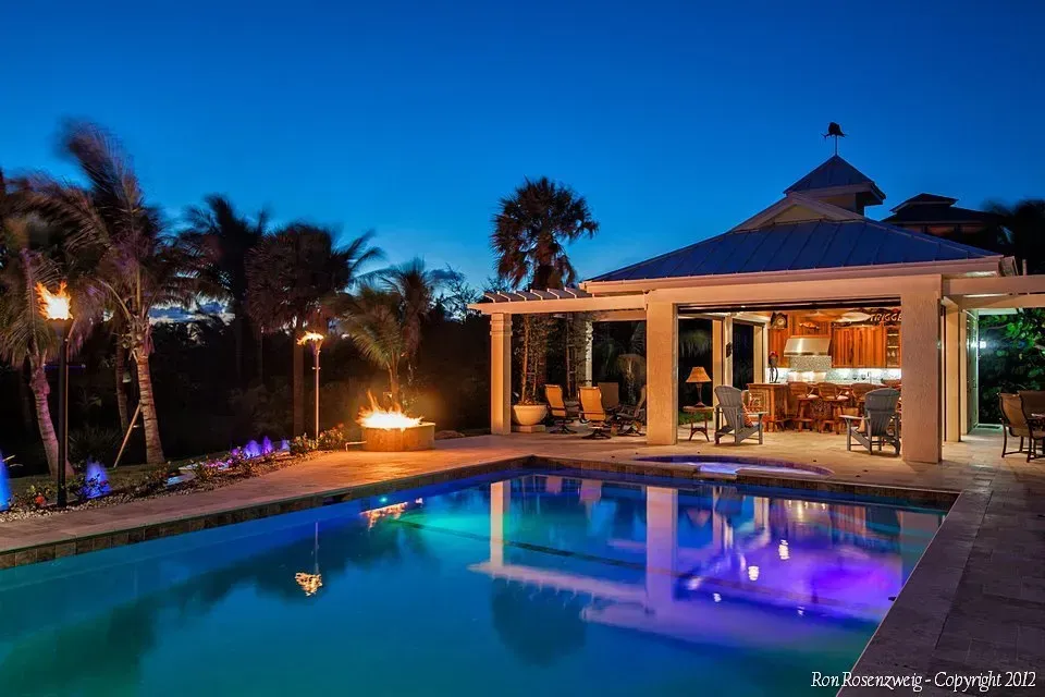 Luxury villa with lit pool at dusk, palm trees and a covered patio glowing blue and orange.